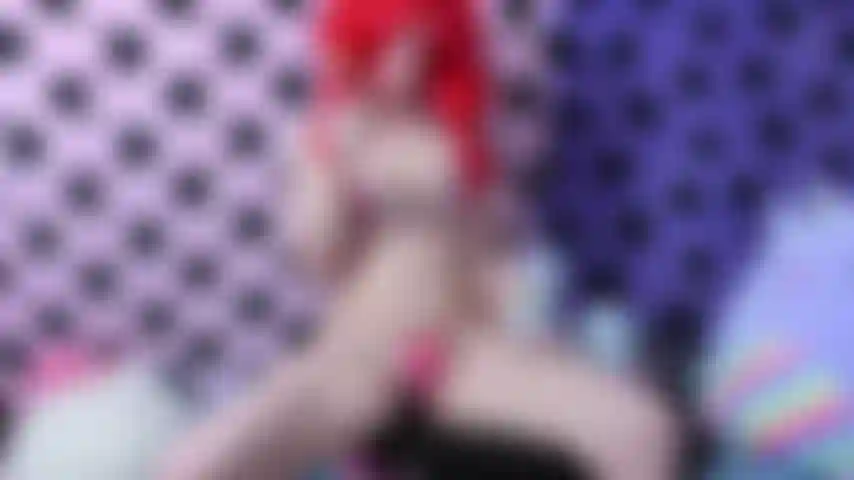 Masturbation mignonne avec un gode en corne de licorne de couleur pastel en bikini coeur et perruque rouge tout en léchant une sucette colorée.